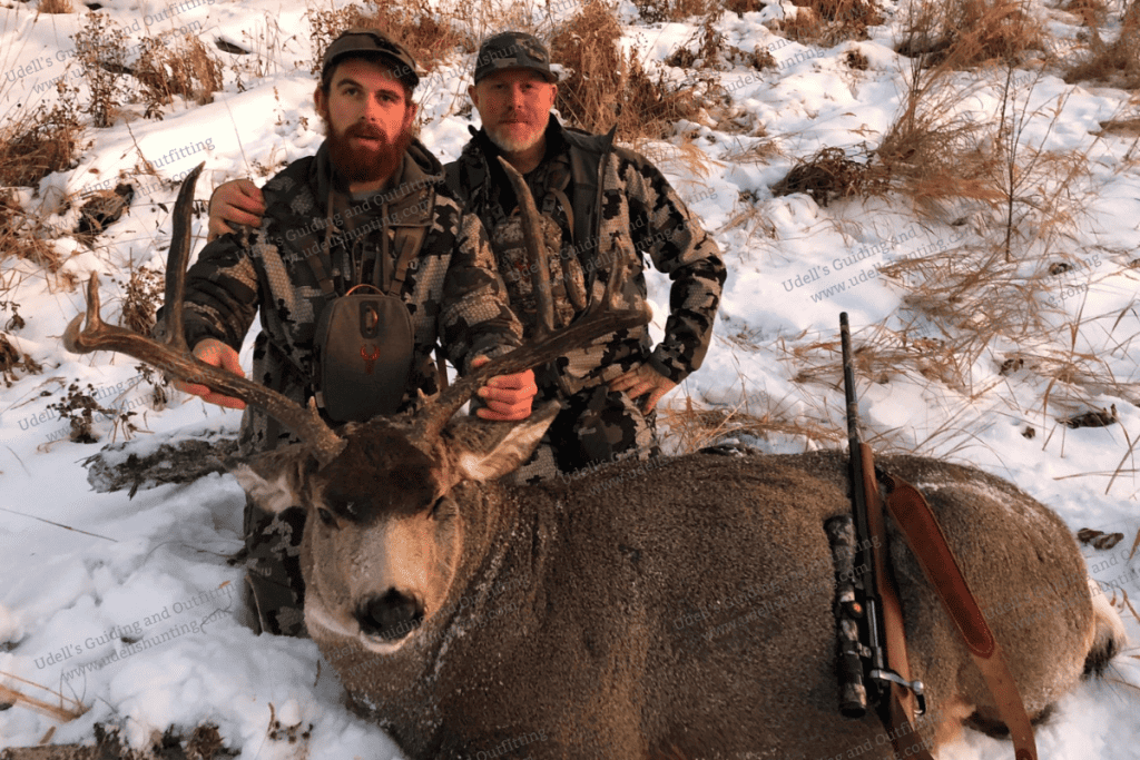 Two men pose with a deer in the snow.